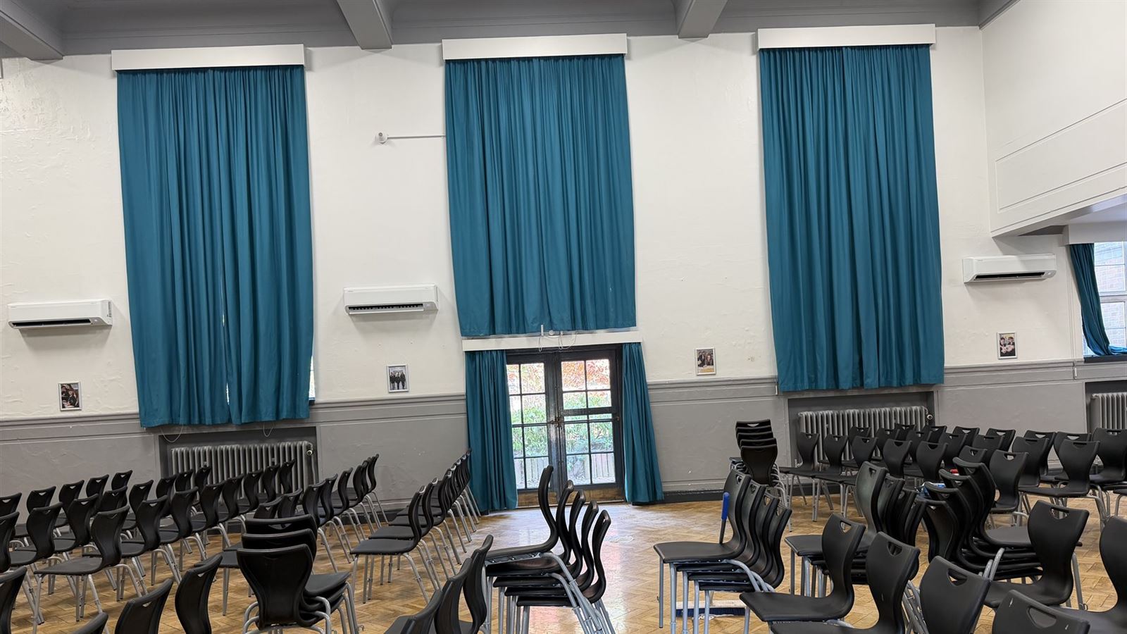 Three Samsung wall-mounted air conditioning units installed high on the wall of a school assembly hall