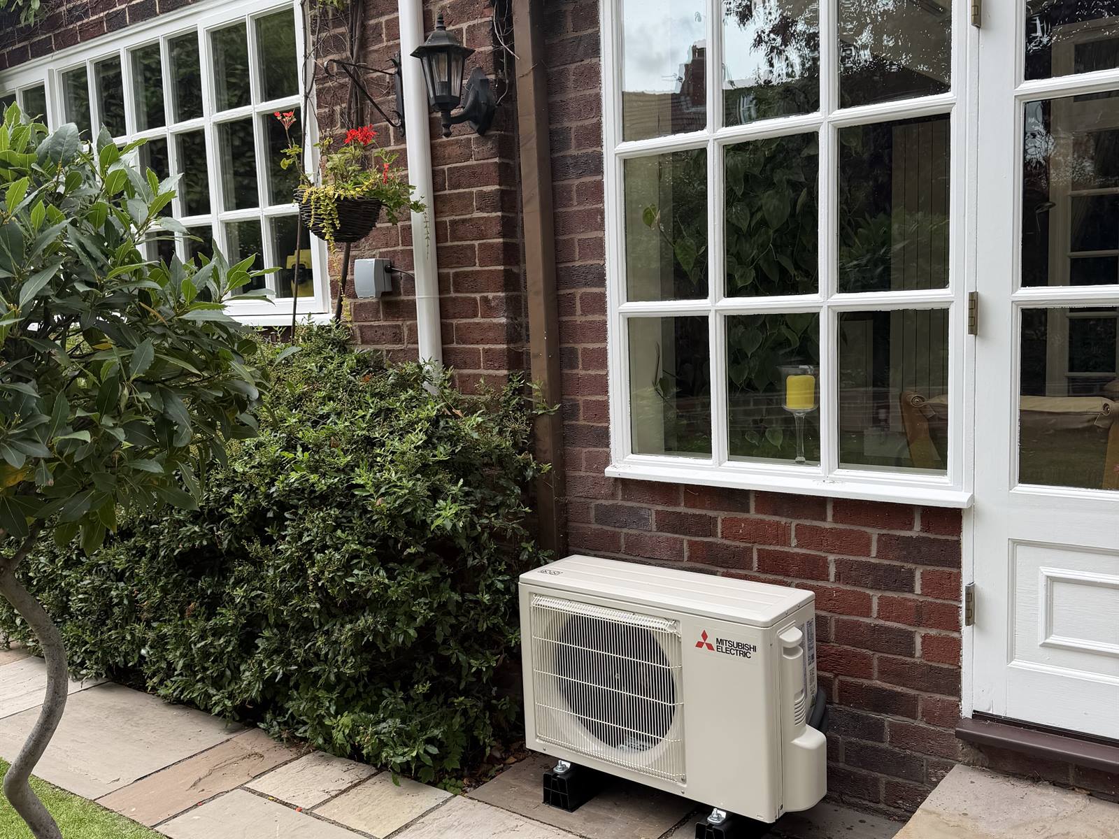 Mitsubishi Electric outdoor condenser unit standing on anti-vibration feet on paving, tucked neatly against the wall beside a door