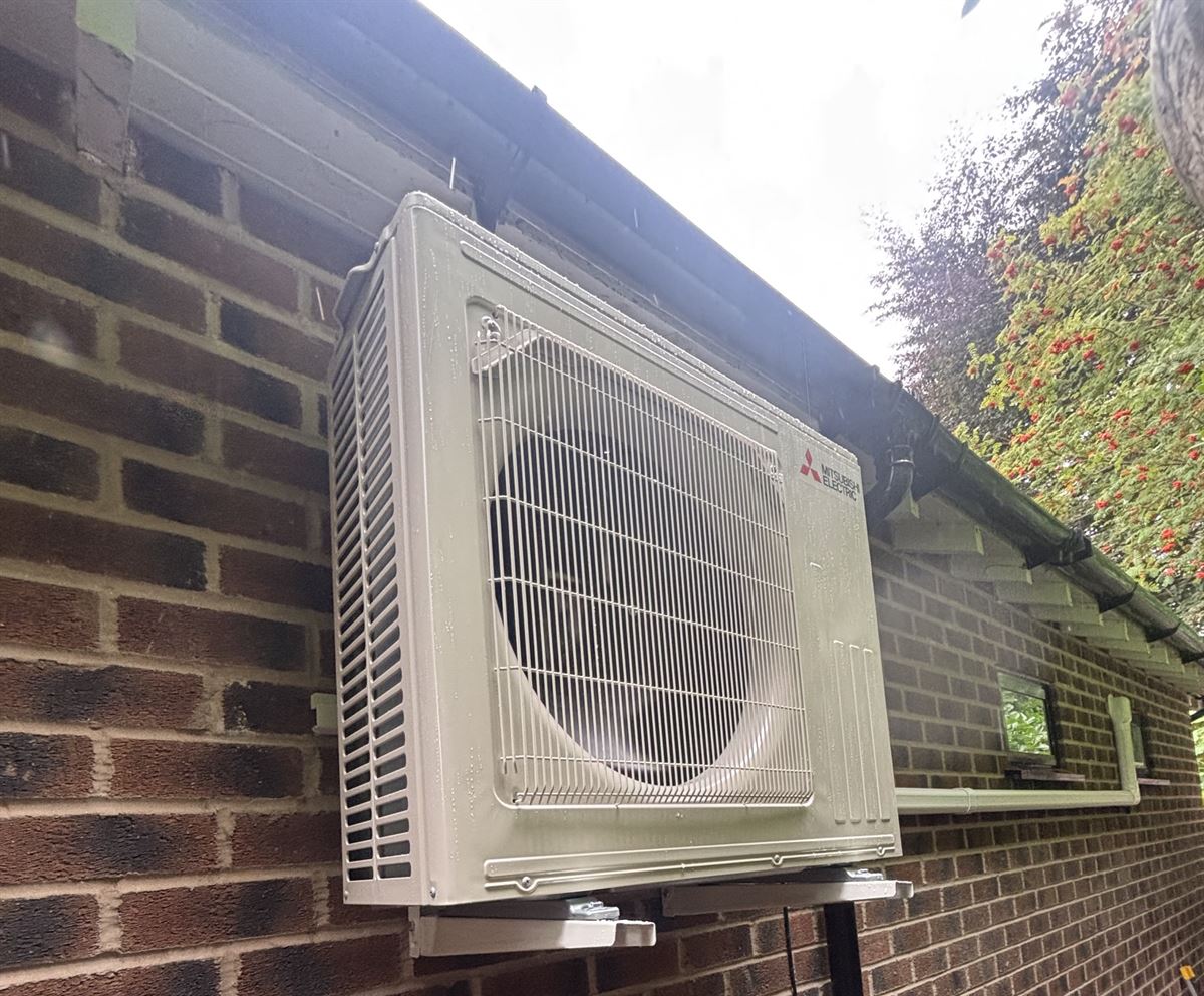 Mitsubishi Electric outdoor unit wall-mounted on brackets at the back of a brick extension, with pipework in white trunking
