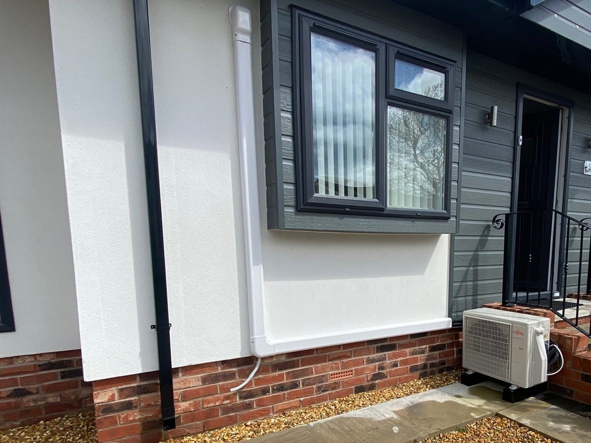 Air conditioning outdoor unit beside a park home on Mossways Park in Wilmslow, with white pipe trunking running down the wall below the window